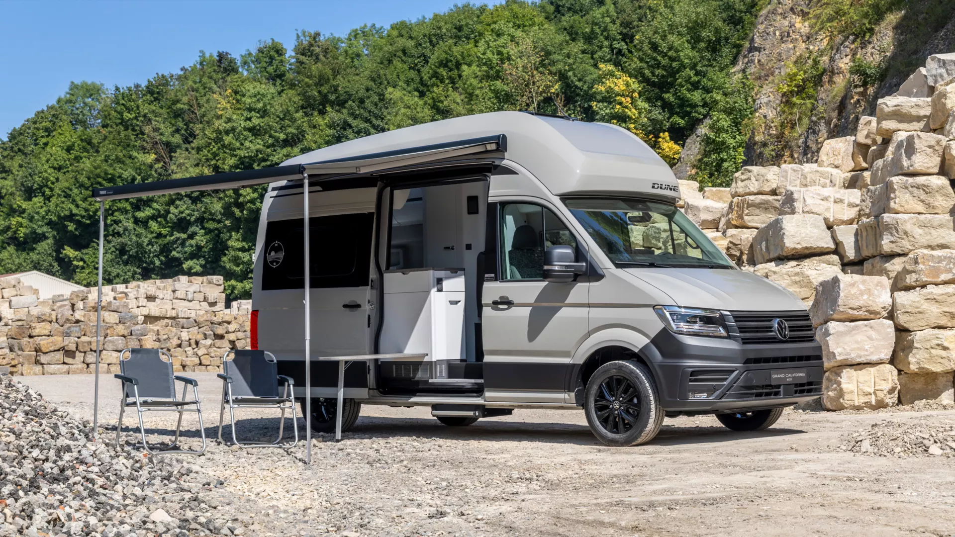 Grand California DUNE steht auf einem steinigen Platz vor einem Hügel mit grünem Bewuchs. Neben dem Wohnmobil sind zwei Campingstühle und ein ausgefahrener Schatten. Das Fahrzeug hat ein erhöhtes Dach und eine Tür, die in den Wohnbereich führt.