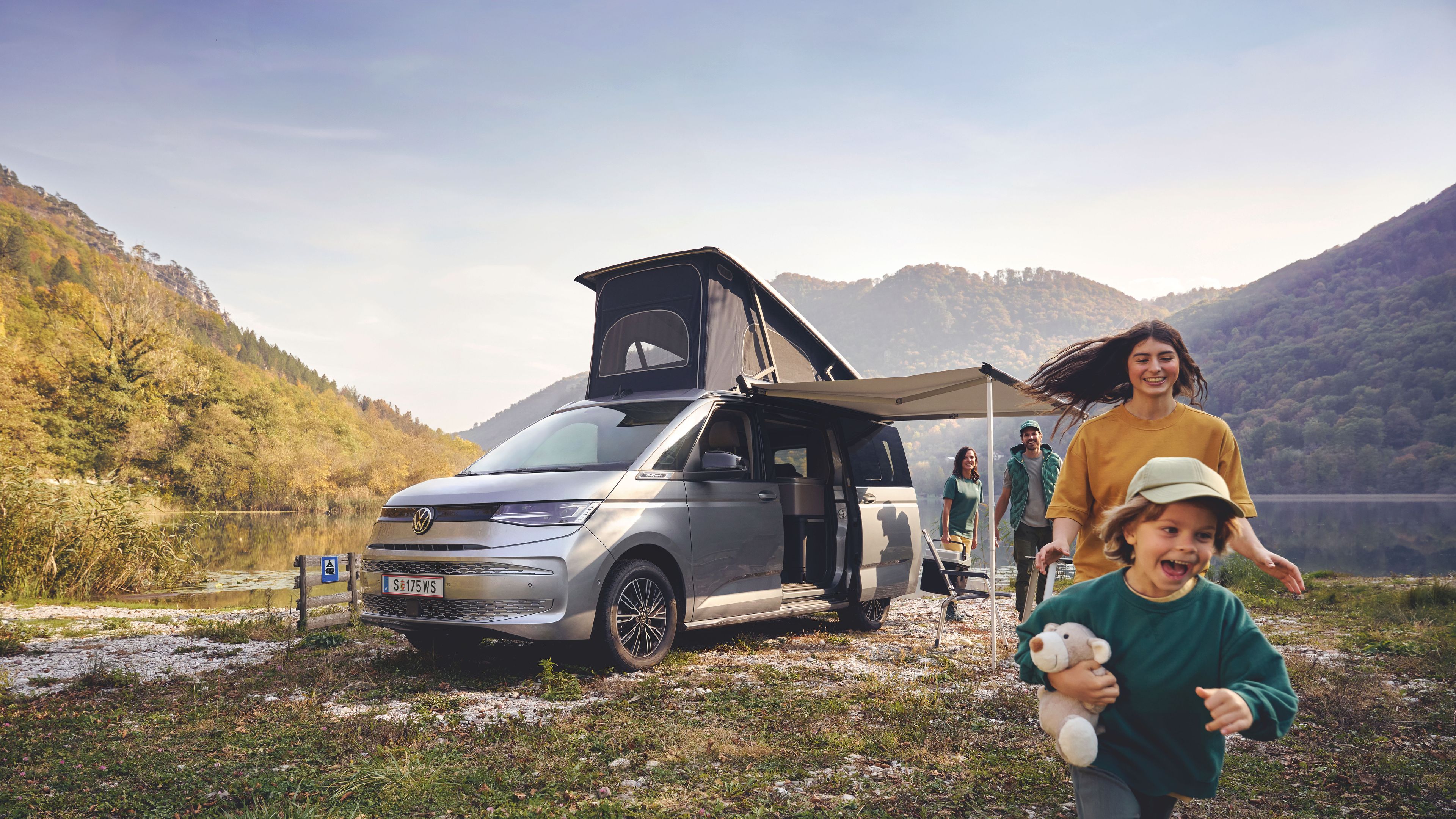 Eine Familie spielt vor ihrem Volkswagen California, welcher an einem See geparkt ist.