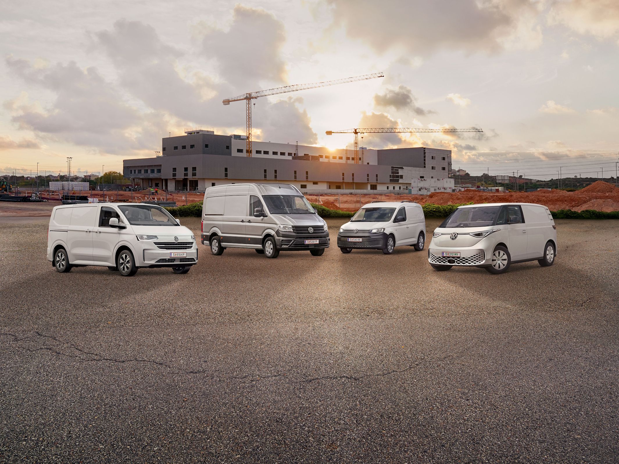 Vier weiße Transporter von Volkswagen Nutzfahrzeuge stehen nebeneinander auf einem leeren Parkplatz vor einem unfertigen Gebäude. Der Himmel ist bewölkt mit einigen Sonnenstrahlen. Ein Baukran ist im Hintergrund sichtbar.
