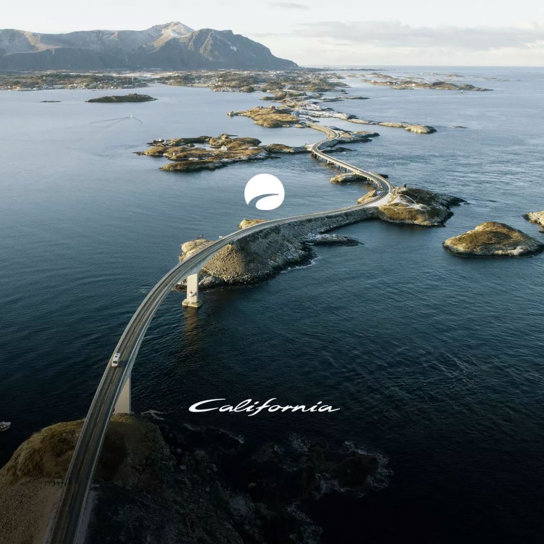 Luftaufnahme einer gebogenen Brücke, die über mehrere Inseln und das Meer führt. Die Landschaft ist karg, mit wenigen Pflanzen und Felsen. Im Hintergrund sind schneebedeckte Berge.