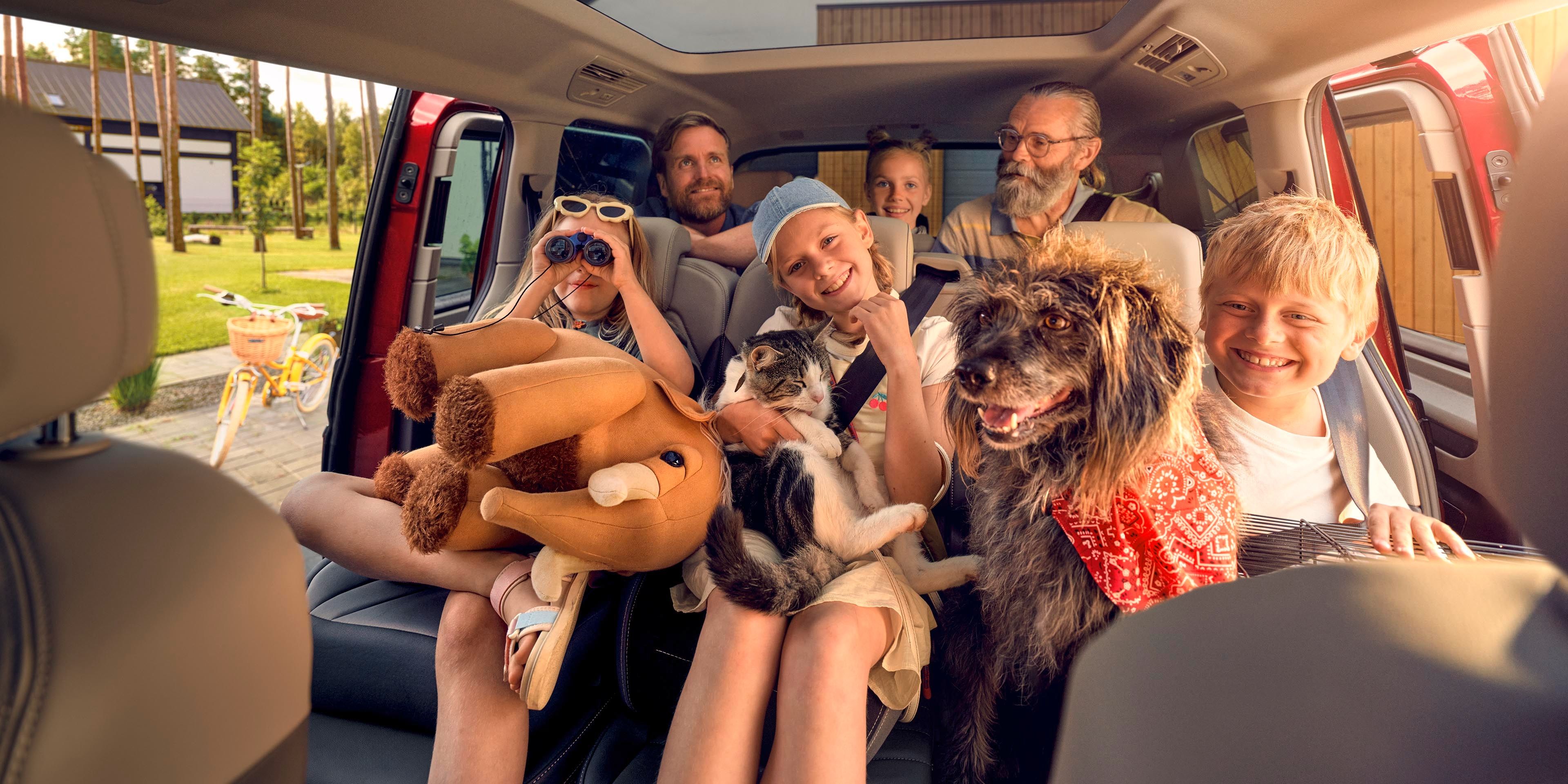 Mehrere Personen sitzen in einem geräumigen Auto, das durch ein großes Panoramadach viel Tageslicht erhält. Ein Hund und eine Katze sind ebenfalls mit im Fahrzeug. Eine Person hat ein großes Plüschtier. Im Hintergrund sind Häuser und Bäume zu sehen.