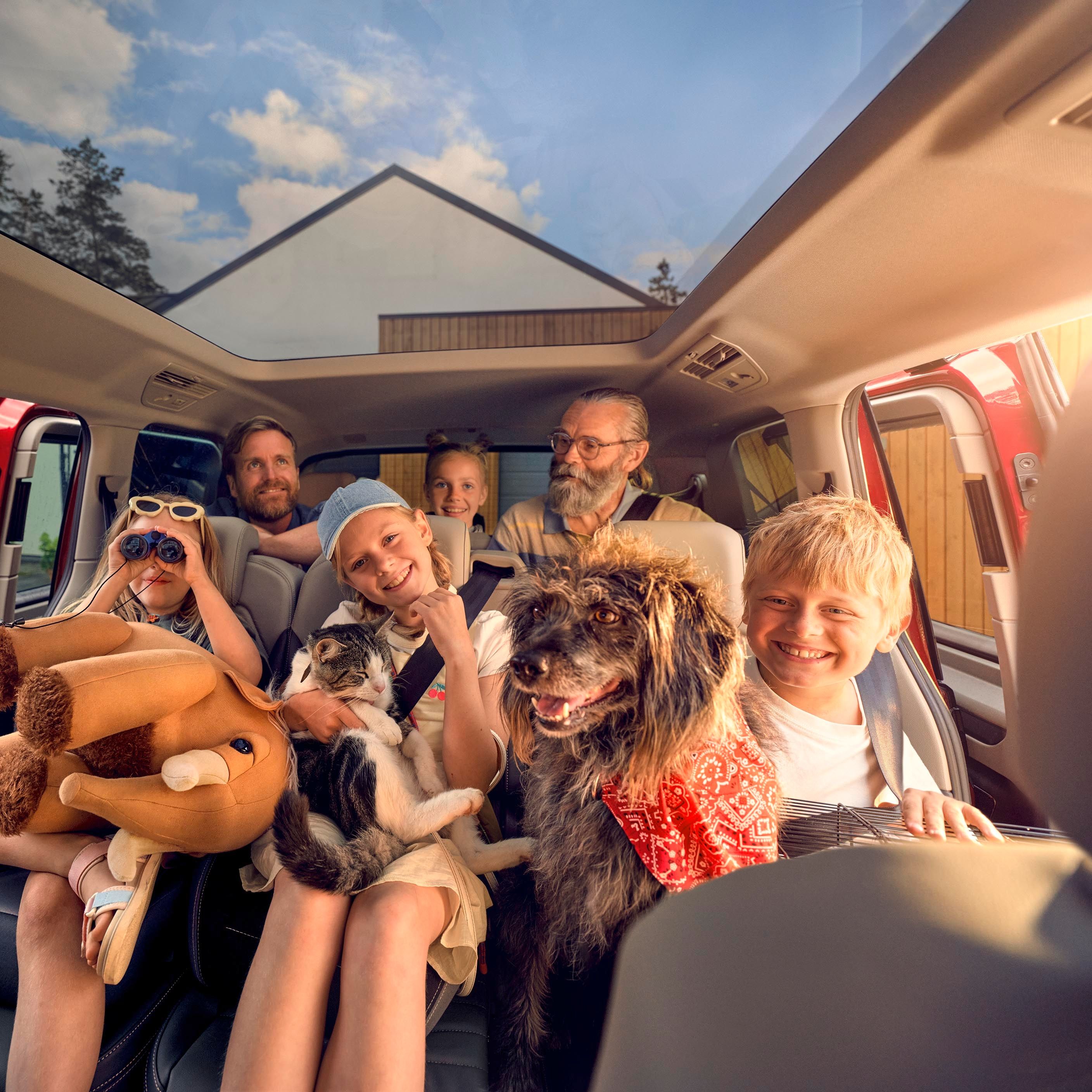 Mehrere Personen sitzen in einem geräumigen Auto, das durch ein großes Panoramadach viel Tageslicht erhält. Ein Hund und eine Katze sind ebenfalls mit im Fahrzeug. Eine Person hat ein großes Plüschtier. Im Hintergrund sind Häuser und Bäume zu sehen.