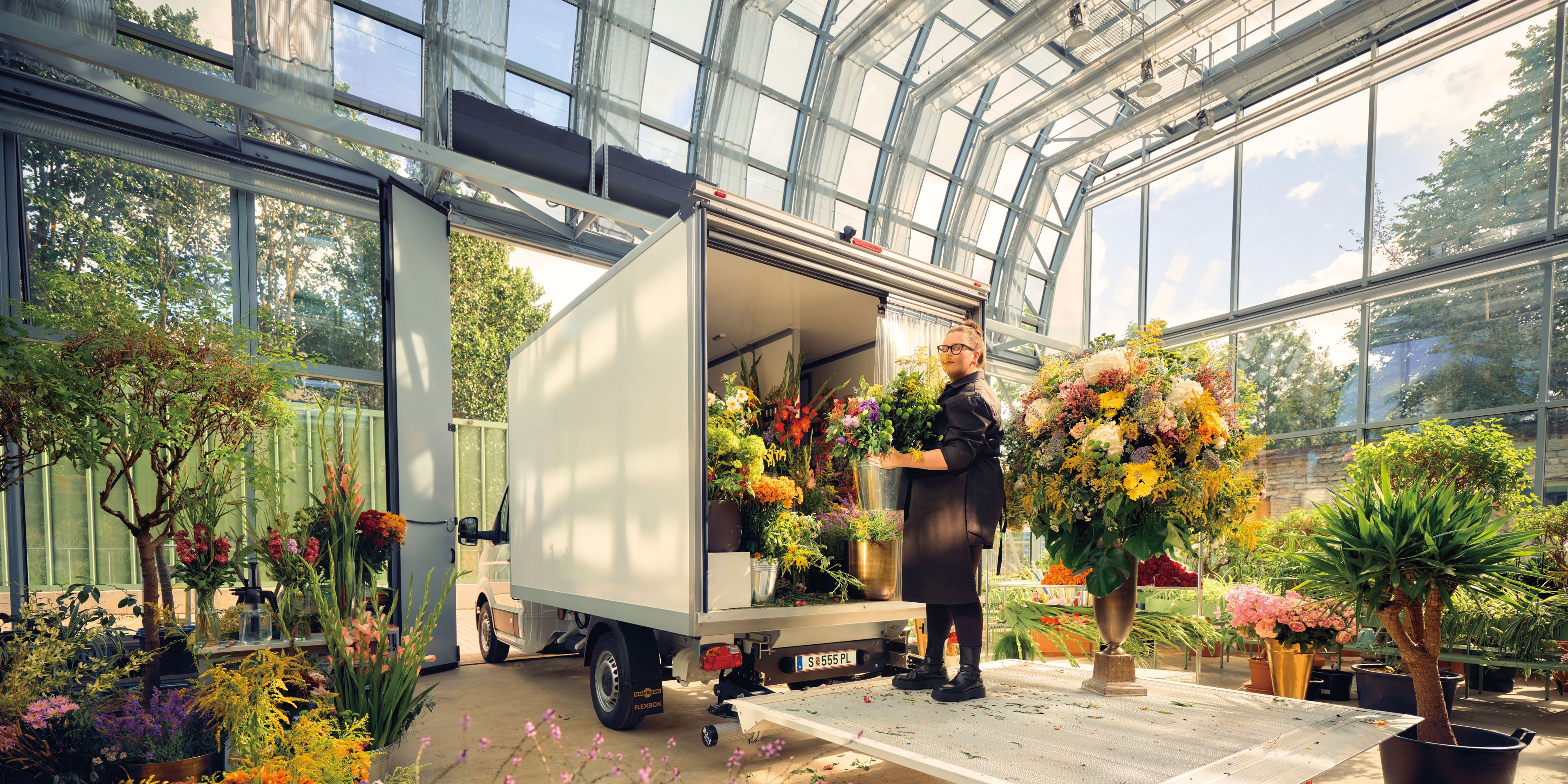 Eine Gärtnerin im Glashaus mit vielen Pflanzen steht auf der Ladefläche eines Volkswagen Transporters.