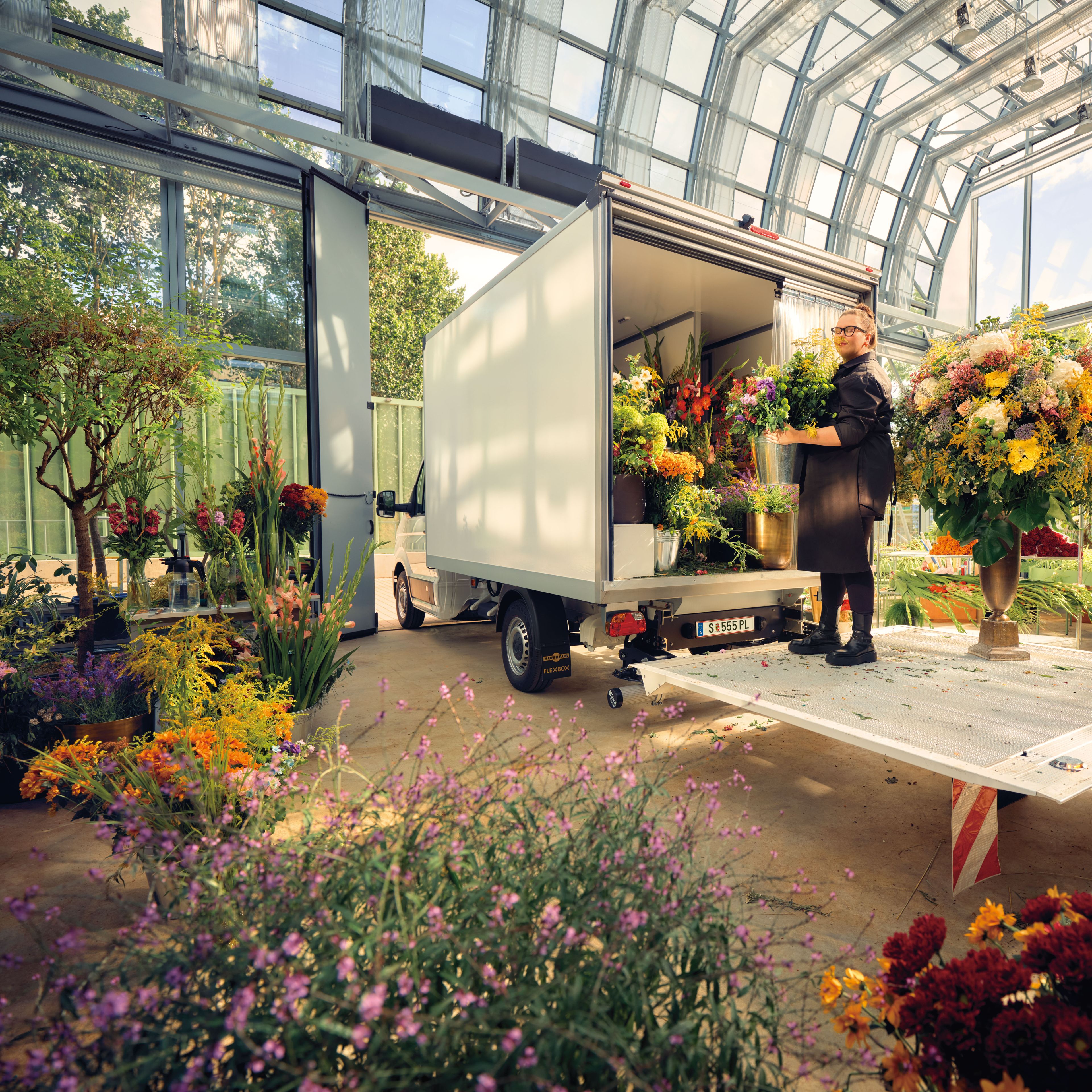 Eine Gärtnerin im Glashaus mit vielen Pflanzen steht auf der Ladefläche eines Volkswagen Transporters.