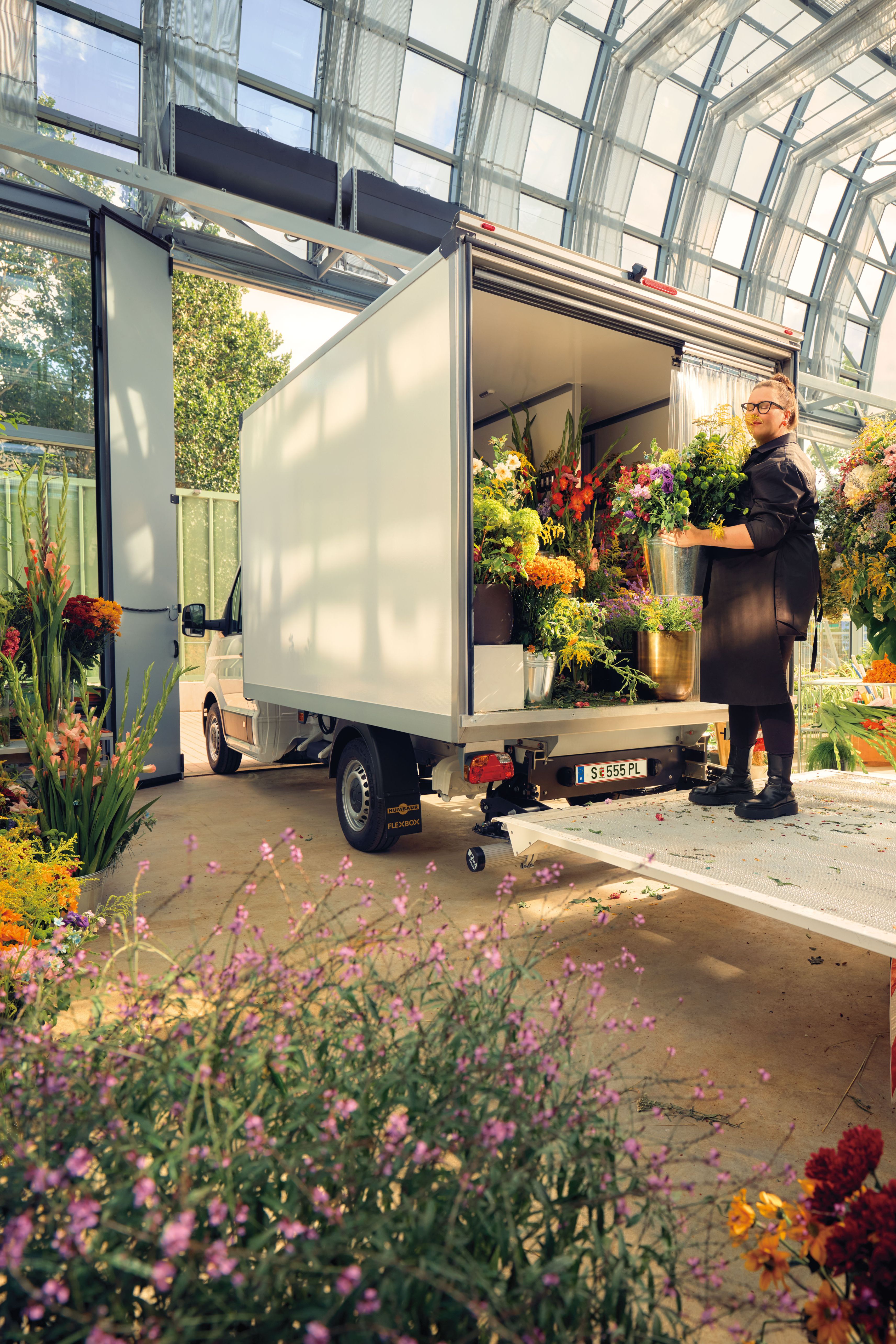 Eine Gärtnerin im Glashaus mit vielen Pflanzen steht auf der Ladefläche eines Volkswagen Transporters.