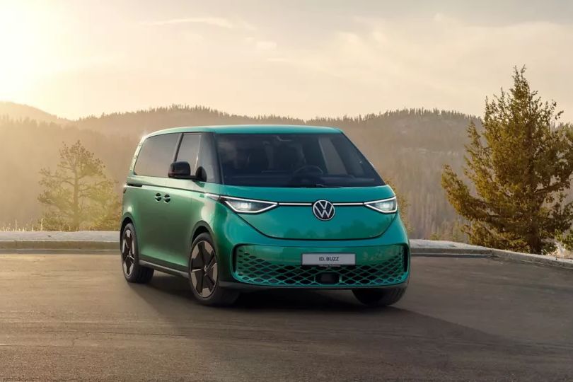 Ein grüner Volkswagen ID. Buzz steht auf einer asphaltierten Fläche, umgeben von Natur und Bergen im Hintergrund bei Sonnenaufgang oder Sonnenuntergang. Der elektrische Kleinbus hat futuristisches Design und moderne Leuchten.