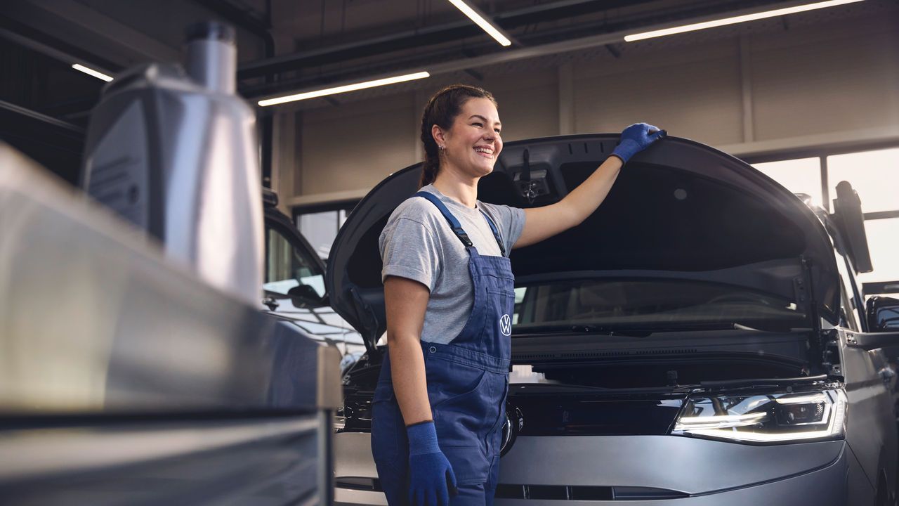 Eine Volkswagen Mechanikerin in blauer Arbeitskleidung steht in einer Volkswagen Werkstatt und inspiziert den Motorraum eines offenen Volkswagen. Sie trägt Handschuhe und stützt sich mit einer Hand auf die Motorhaube.