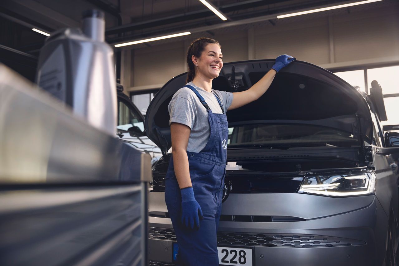 Eine Volkswagen Mechanikerin in blauer Arbeitskleidung steht in einer Volkswagen Werkstatt und inspiziert den Motorraum eines offenen Volkswagen. Sie trägt Handschuhe und stützt sich mit einer Hand auf die Motorhaube.