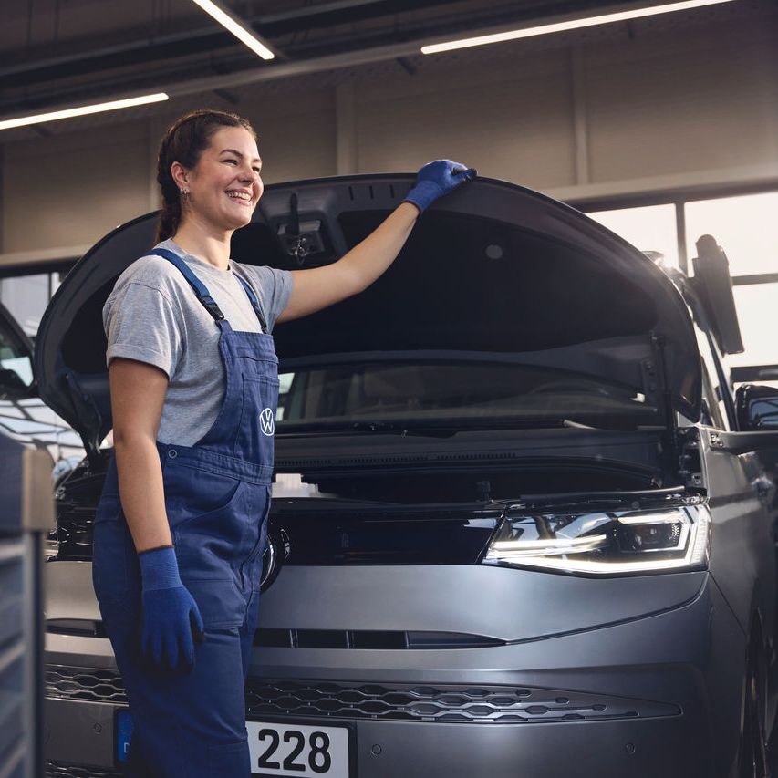 Eine Volkswagen Mechanikerin in blauer Arbeitskleidung steht in einer Volkswagen Werkstatt und inspiziert den Motorraum eines offenen Volkswagen. Sie trägt Handschuhe und stützt sich mit einer Hand auf die Motorhaube.
