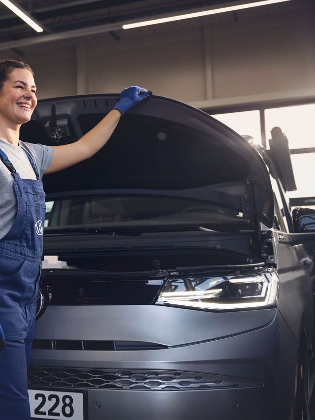Eine Volkswagen Mechanikerin in blauer Arbeitskleidung steht in einer Volkswagen Werkstatt und inspiziert den Motorraum eines offenen Volkswagen. Sie trägt Handschuhe und stützt sich mit einer Hand auf die Motorhaube.