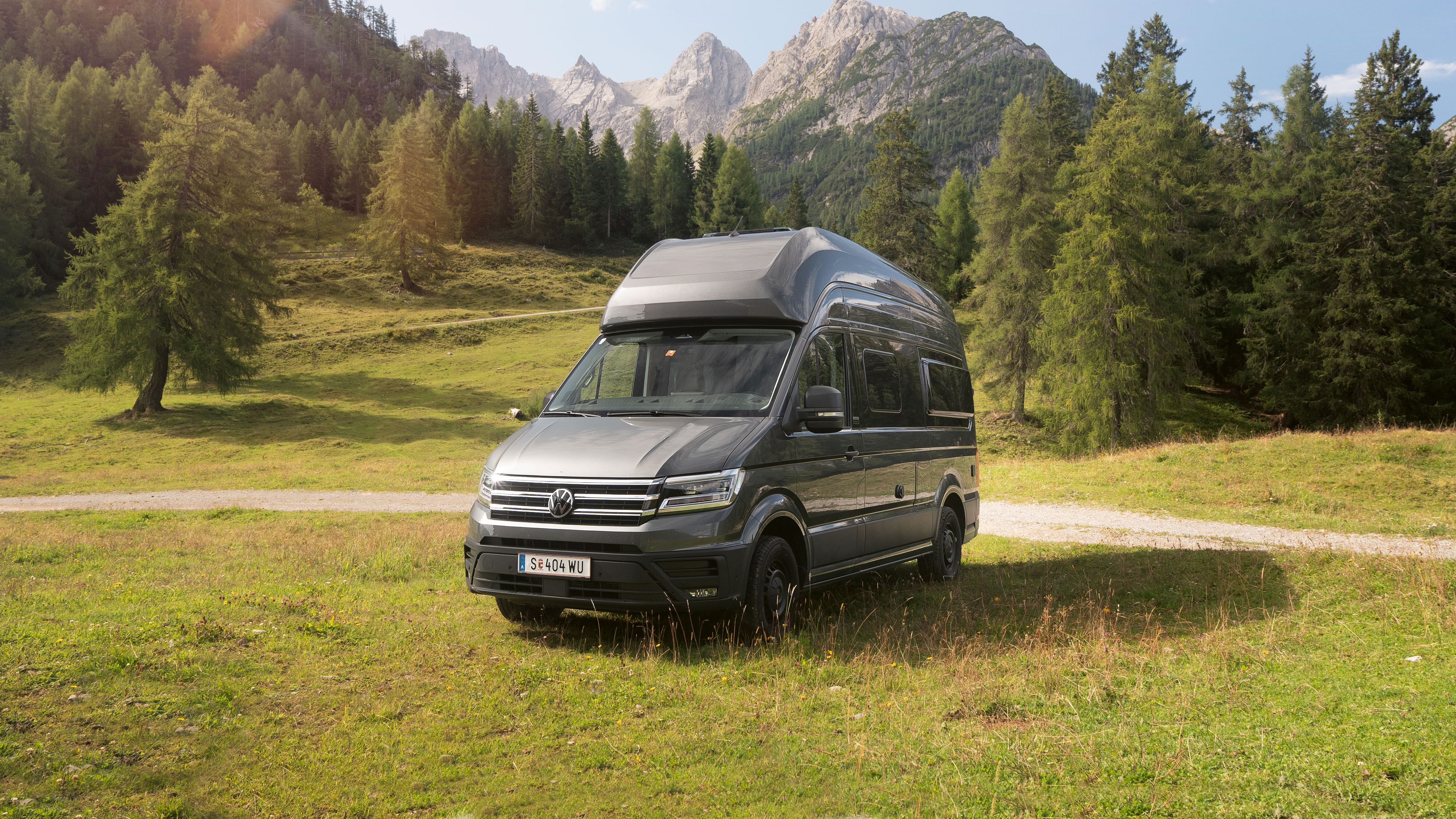 Ein silberner Wohnmobil-Van steht auf einer grünen Wiese vor einer malerischen Berglandschaft mit Bäumen und blauem Himmel.