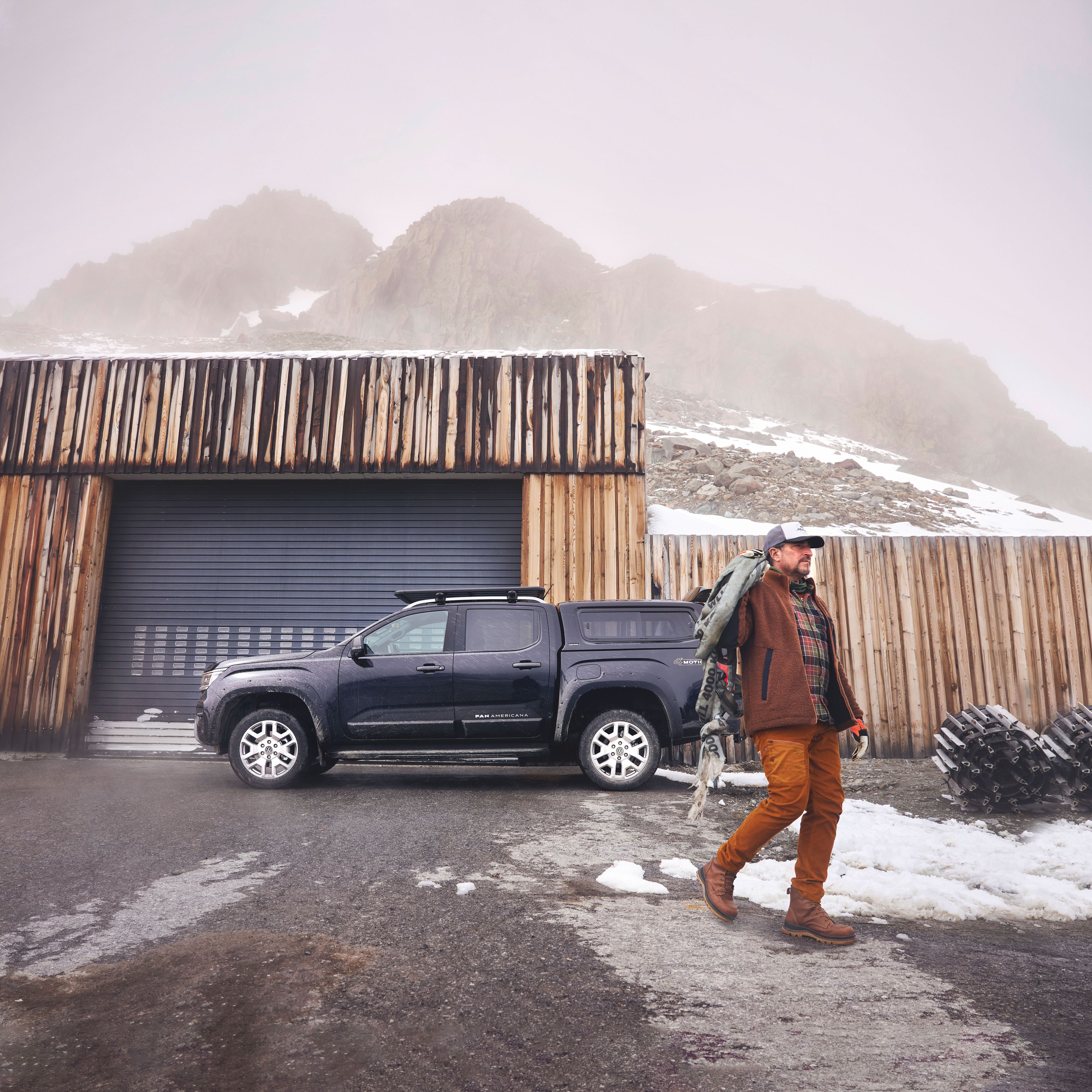 Ein Mann in Outdoor-Kleidung geht auf einem nassen, teils verschneiten Platz. Hinter ihm steht ein dunkler Volkswagen Amarok vor einer hölzernen Garage, im Hintergrund ragen nebelverhangene Felsen auf.