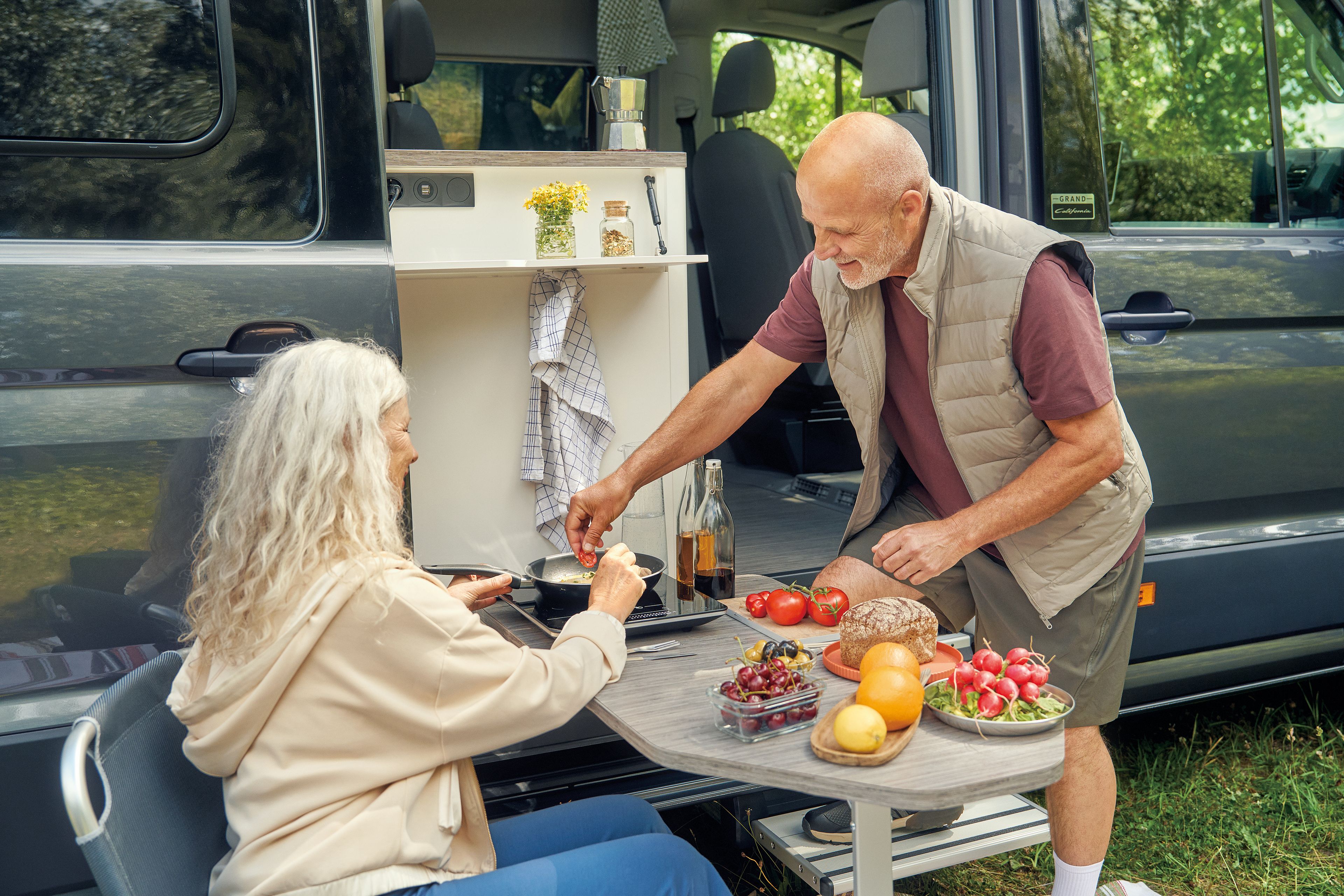 Ein älteres Paar sitzt vor einem Wohnmobil und bereitet gemeinsam eine Mahlzeit zu. Auf dem Tisch befinden sich verschiedene frische Lebensmittel, darunter Tomaten, Radieschen und Brot. Die Umgebung ist grün und einladend.