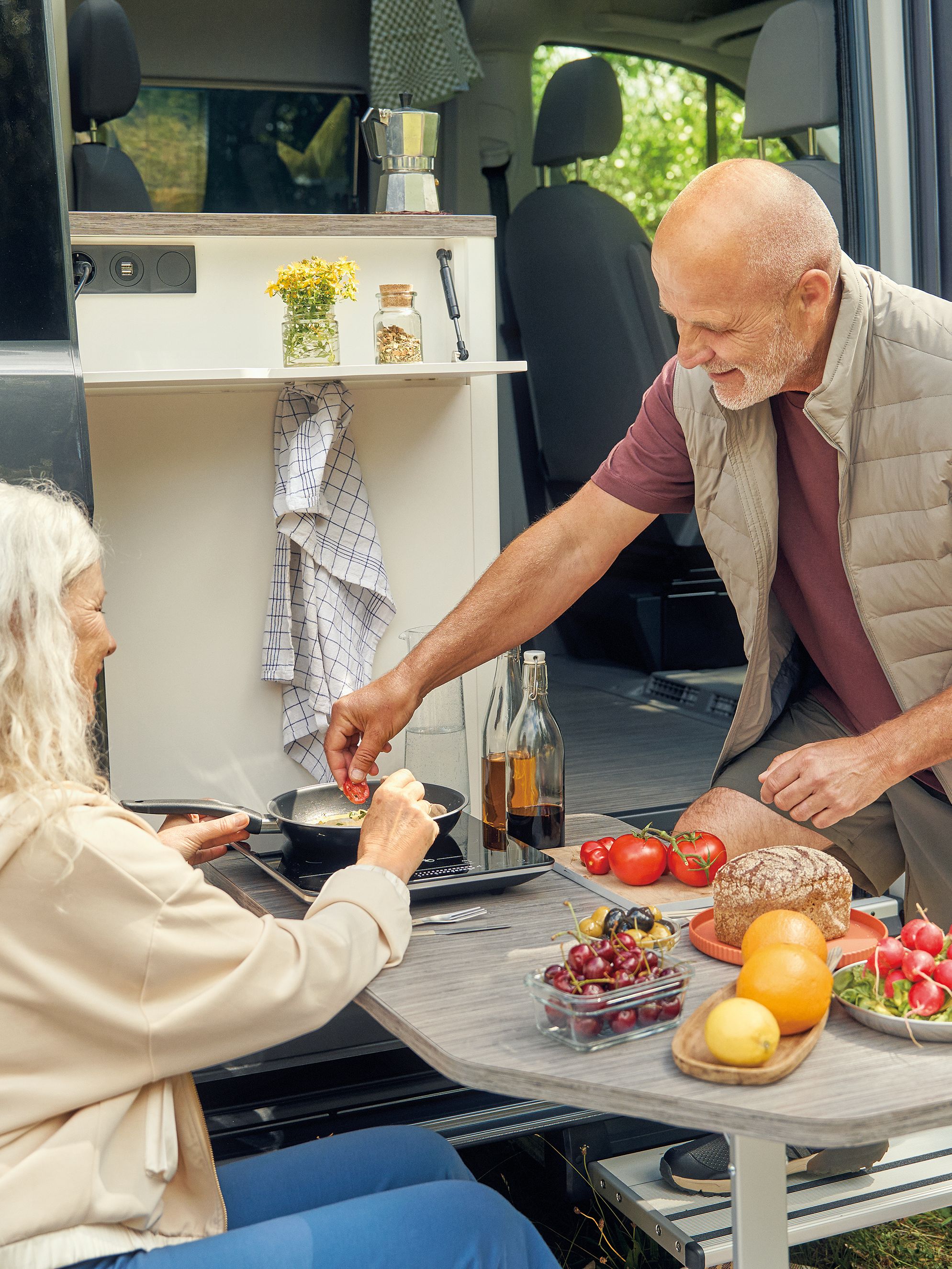 Ein älteres Paar sitzt vor einem Wohnmobil und bereitet gemeinsam eine Mahlzeit zu. Auf dem Tisch befinden sich verschiedene frische Lebensmittel, darunter Tomaten, Radieschen und Brot. Die Umgebung ist grün und einladend.