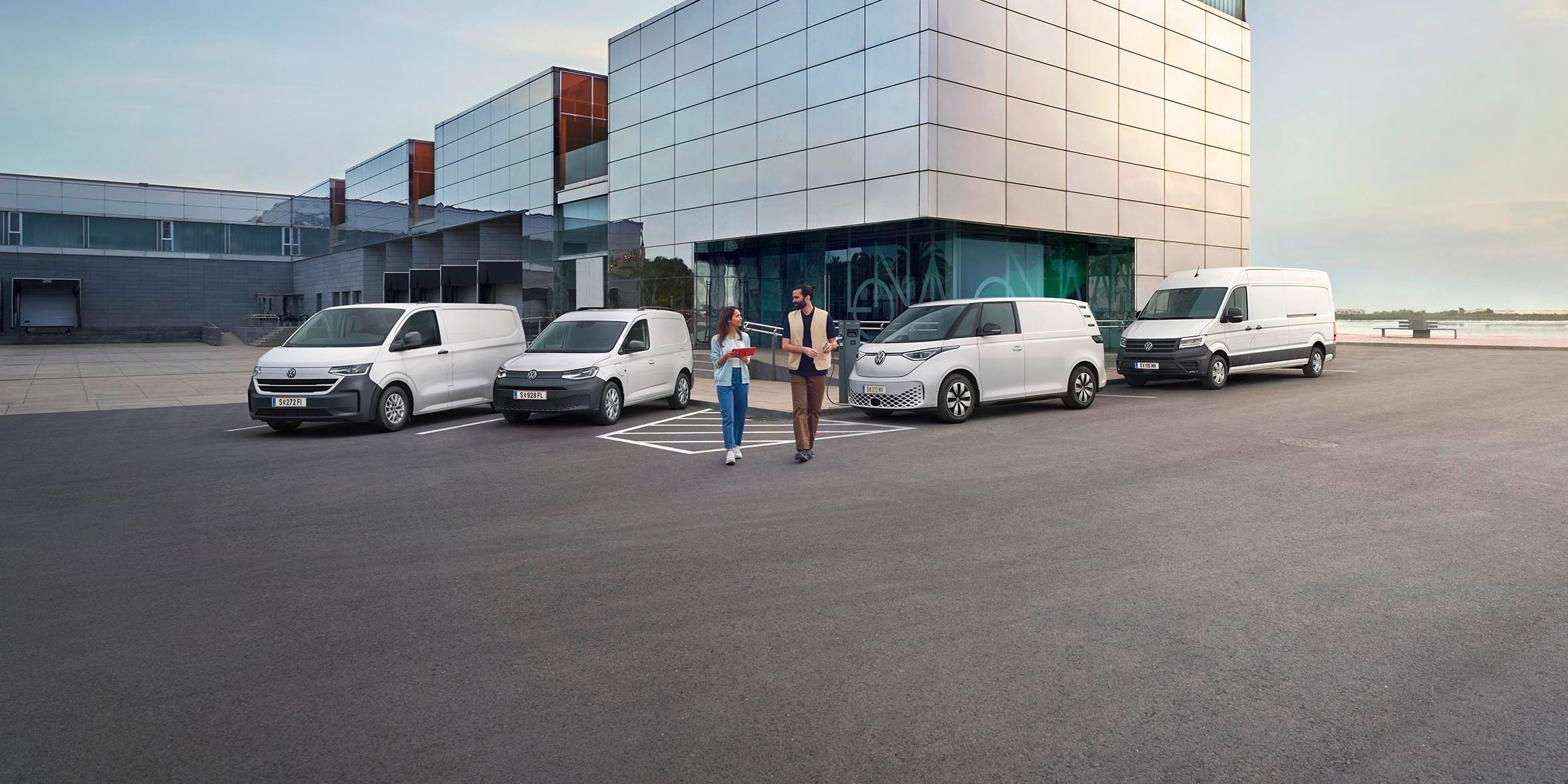 Ein modernes Lagergebäude mit Verladebereichen und mehreren geparkten Transportern von Volkswagen Nutzfahrzeuge. Zwei Personen stehen vor dem Gebäude und unterhalten sich. Der Himmel ist teilweise bewölkt, und es gibt einen Baum in der Ferne.