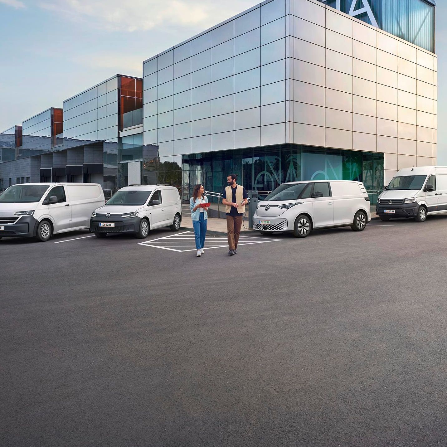 Ein modernes Lagergebäude mit Verladebereichen und mehreren geparkten Transportern von Volkswagen Nutzfahrzeuge. Zwei Personen stehen vor dem Gebäude und unterhalten sich. Der Himmel ist teilweise bewölkt, und es gibt einen Baum in der Ferne.