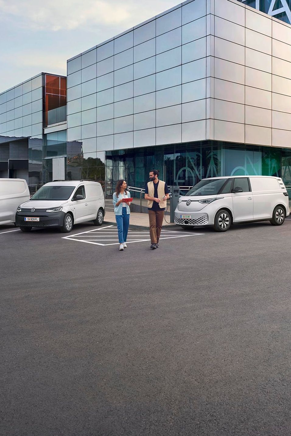 Ein modernes Lagergebäude mit Verladebereichen und mehreren geparkten Transportern von Volkswagen Nutzfahrzeuge. Zwei Personen stehen vor dem Gebäude und unterhalten sich. Der Himmel ist teilweise bewölkt, und es gibt einen Baum in der Ferne.