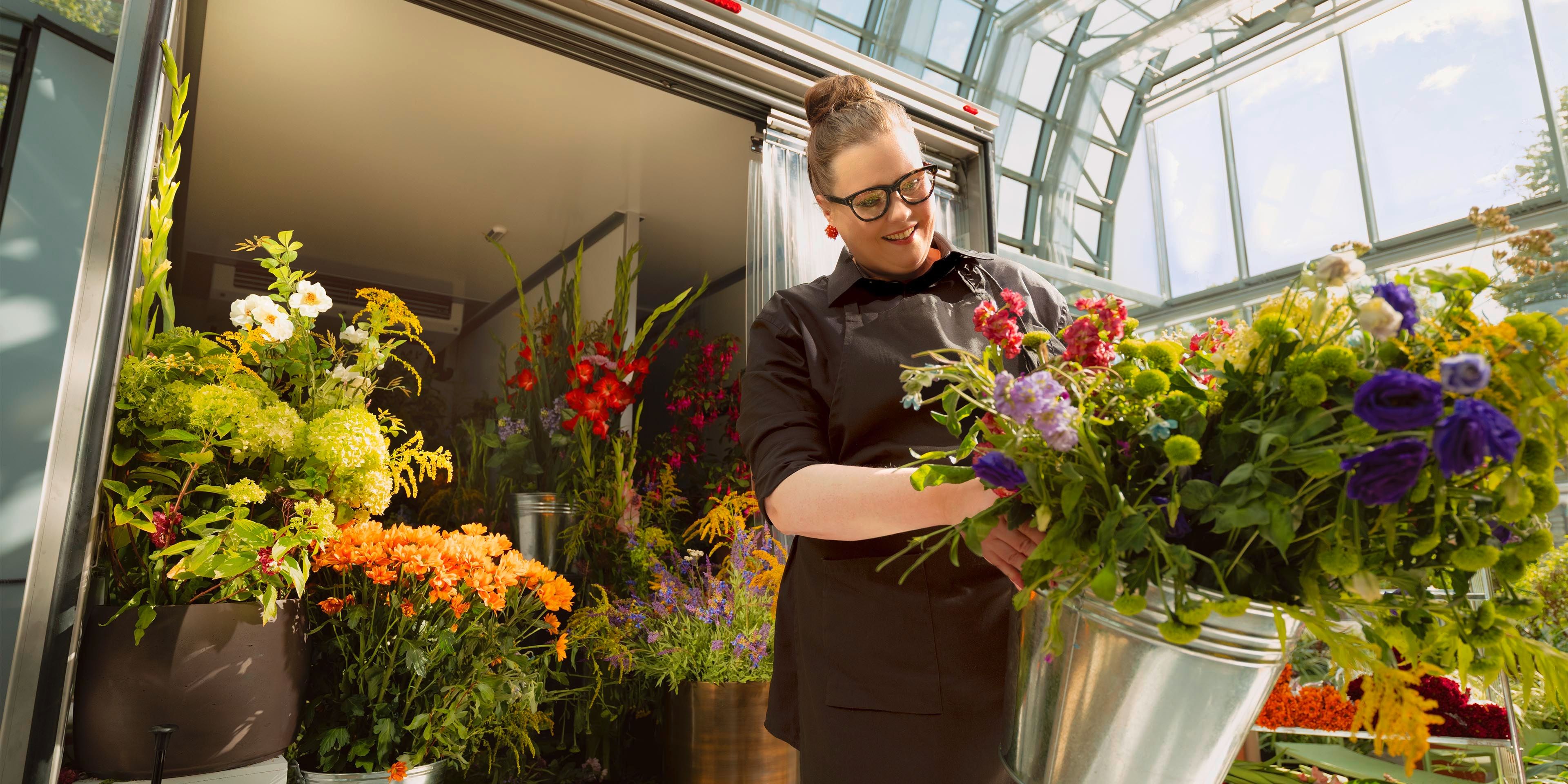 Eine Person steht in einem Blumenladen, umgeben von einer Vielzahl von bunten Blumenarrangements. Sie hält einen silbernen Eimer voller frischer Blumen. Der Raum hat große Fenster, die viel Tageslicht hereinlassen.