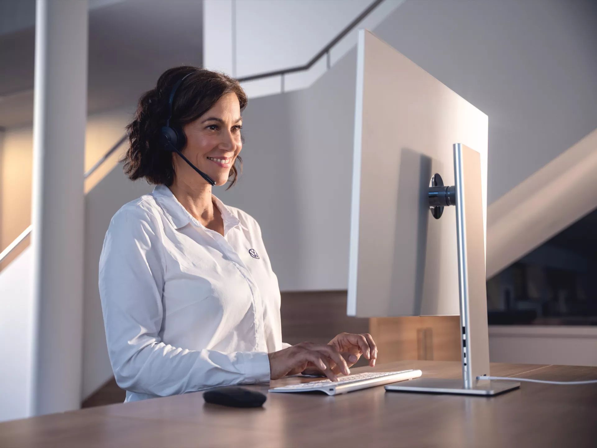 Frau mittleren Alters mit Headset sitzt an einem Schreibtisch und tippt lächelnd auf einer Tastatur vor einem großen Monitor. Im Hintergrund ist eine moderne, helle Büro- oder Homeoffice-Umgebung mit Treppe zu sehen.