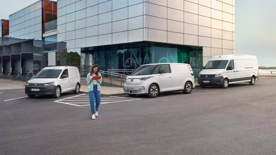 Eine Frau mit einem Tablet steht vor einem Gebäude mit Glasfront. Im Hintergrund sind drei verschiedene Lieferwagen geparkt. Sie sind weiß und unterschiedlich groß.