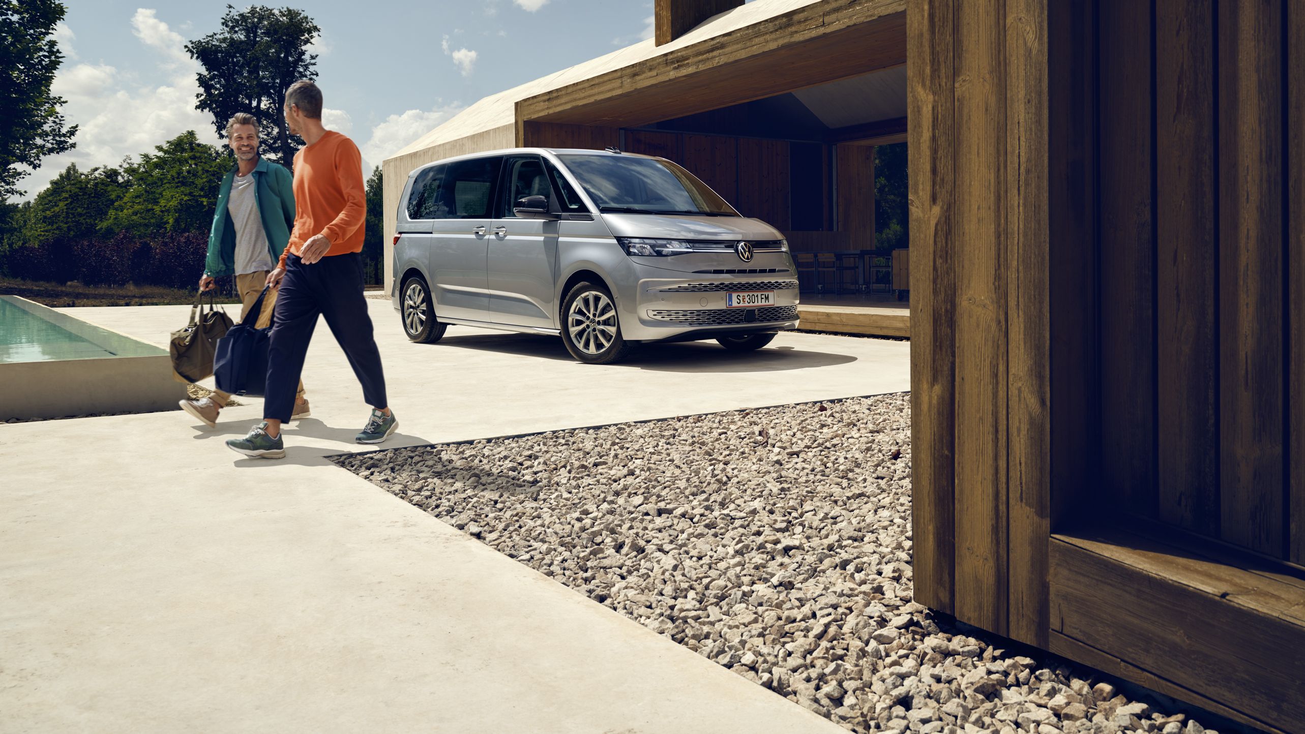 Ein Volkswagen Multivan in Silber parkt vor einem Haus mit Pool, zwei Männer mit Sporttaschen bewegen sich von dem Fahrzeug fort.
