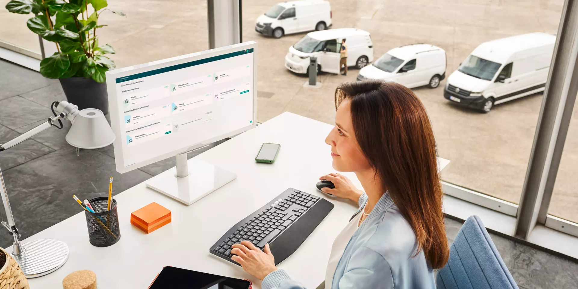Eine Frau sitzt an einem Schreibtisch vor einem Computer in einem modernen Büro mit großen Fenstern. Draußen sind mehrere weiße Lieferwagen zu sehen. Auf dem Schreibtisch liegen eine Wasserflasche, ein Tablet und Schreibutensilien.