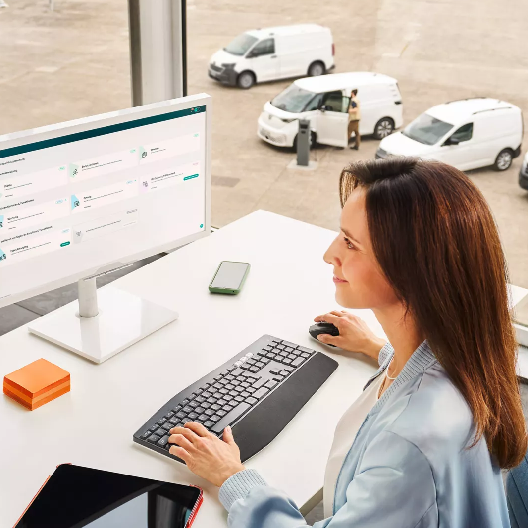 Eine Frau sitzt an einem Schreibtisch vor einem Computer in einem modernen Büro mit großen Fenstern. Draußen sind mehrere weiße Lieferwagen zu sehen. Auf dem Schreibtisch liegen eine Wasserflasche, ein Tablet und Schreibutensilien.