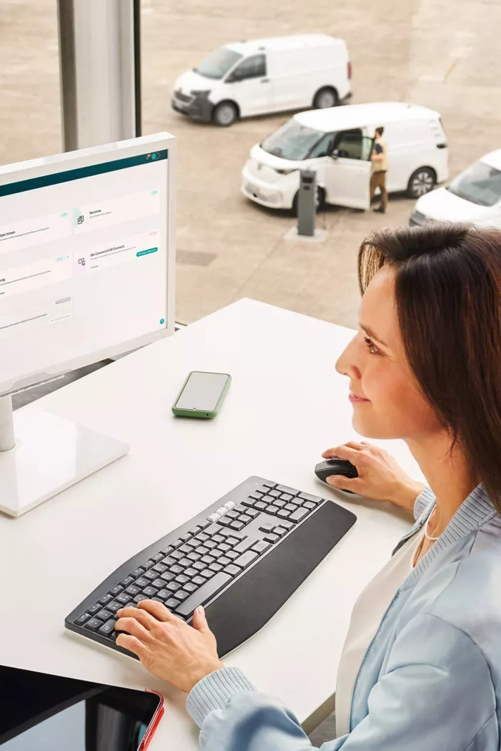 Eine Frau sitzt an einem Schreibtisch vor einem Computer in einem modernen Büro mit großen Fenstern. Draußen sind mehrere weiße Lieferwagen zu sehen. Auf dem Schreibtisch liegen eine Wasserflasche, ein Tablet und Schreibutensilien.