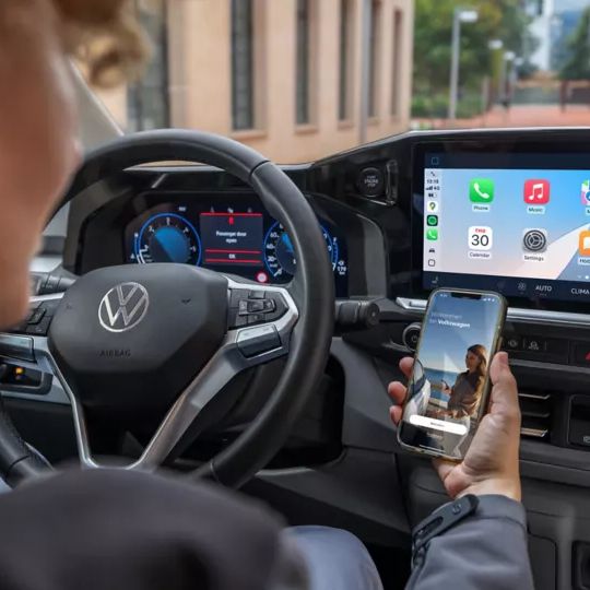 Eine Person sitzt in einem Auto und hält ein Smartphone in der Hand, während auf dem großen Display des Autos verschiedene Apps angezeigt werden. Das Lenkrad zeigt das VW-Logo.