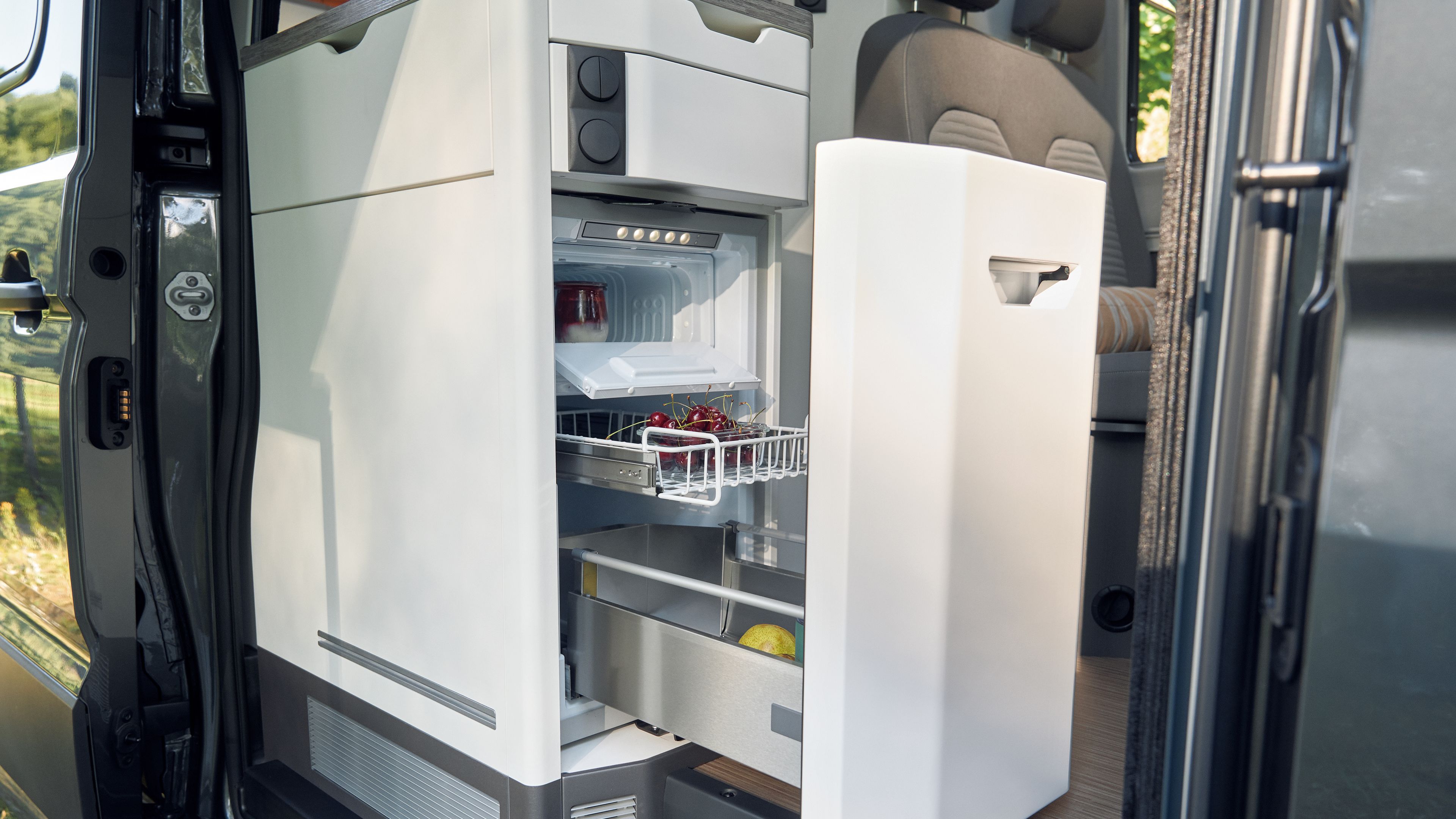 Ein geöffneter Kühlschrank in einem Camper-Van mit mehreren Fächern, in denen Lebensmittel lagern. Einige Getränkedosen sind im oberen Fach sichtbar und Früchte befinden sich im Drahtkorb.