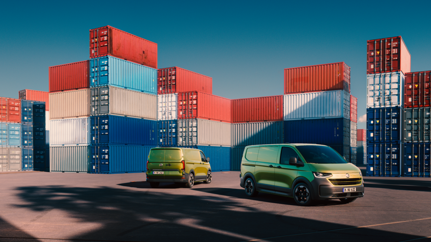 Zwei grüne Volkswagen Transporter vor gestapelten Containern in einem Hafen.