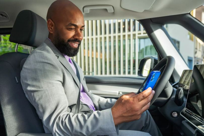 Ein Mann in einem grauen Anzug sitzt in einem Auto und schaut auf sein Smartphone. Sein Gesicht ist unscharf gemacht. Im Hintergrund sind moderne Gebäude zu sehen.