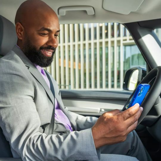 Ein Mann in einem grauen Anzug sitzt in einem Auto und schaut auf sein Smartphone. Sein Gesicht ist unscharf gemacht. Im Hintergrund sind moderne Gebäude zu sehen.