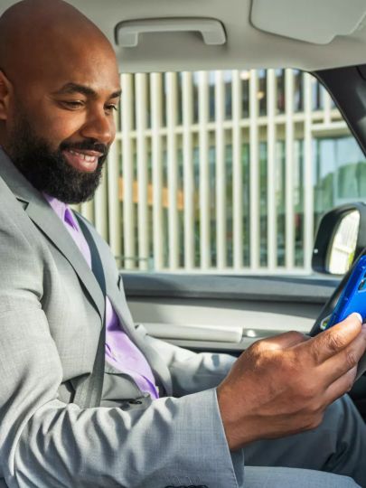 Ein Mann in einem grauen Anzug sitzt in einem Auto und schaut auf sein Smartphone. Sein Gesicht ist unscharf gemacht. Im Hintergrund sind moderne Gebäude zu sehen.