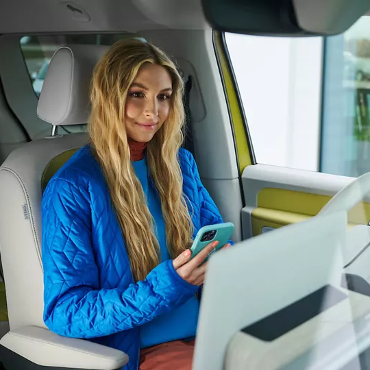 Junge Frau mit langen blonden Haaren sitzt auf dem Fahrersitz eines modernen Autos und hält ein Smartphone in der Hand, während sie lächelnd zur Kamera blickt. Innenraum und Sitze wirken hell und minimalistisch. Rechts im Bild ist unscharf ein rundes Logo auf der Autoscheibe zu sehen.