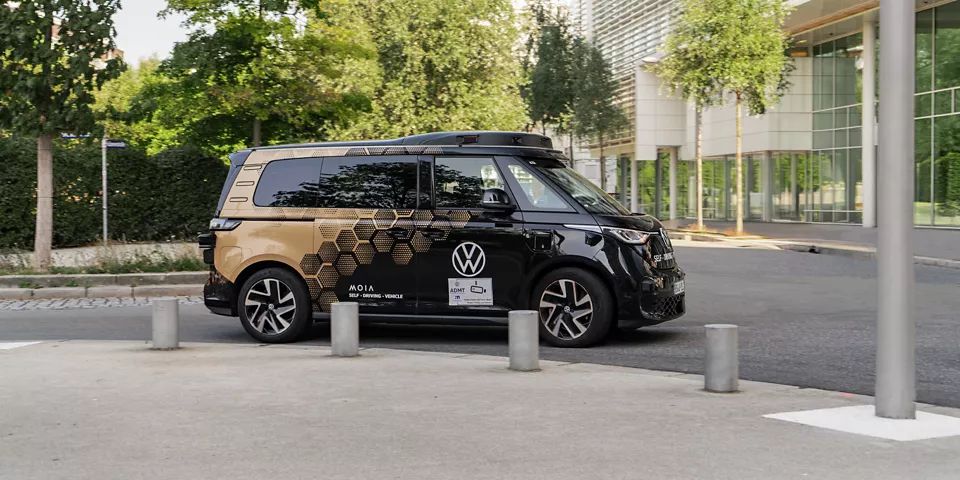 Ein schwarzer Volkswagen-Campervan mit goldenen und schwarzen Wabenmustern parkt auf einer städtischen Straße. Umgeben von modernen Gebäuden und Bäumen. Vor dem Fahrzeug sind mehrere graue Poller sichtbar.