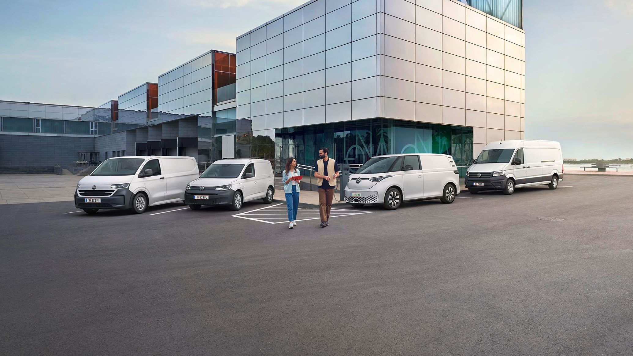 Ein modernes Lagergebäude mit Verladebereichen und mehreren geparkten Transportern von Volkswagen Nutzfahrzeuge. Zwei Personen stehen vor dem Gebäude und unterhalten sich. Der Himmel ist teilweise bewölkt, und es gibt einen Baum in der Ferne.