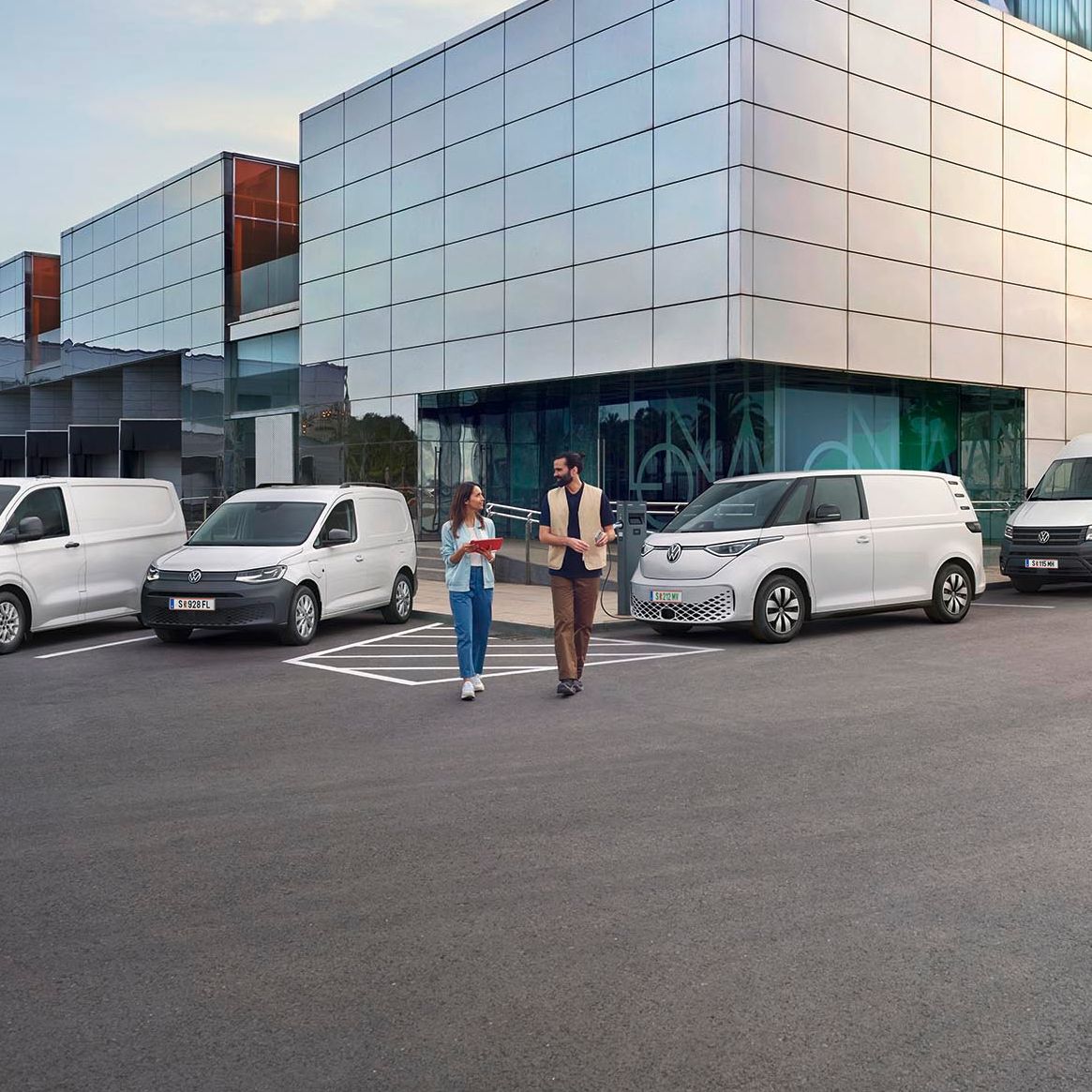 Ein modernes Lagergebäude mit Verladebereichen und mehreren geparkten Transportern von Volkswagen Nutzfahrzeuge. Zwei Personen stehen vor dem Gebäude und unterhalten sich. Der Himmel ist teilweise bewölkt, und es gibt einen Baum in der Ferne.