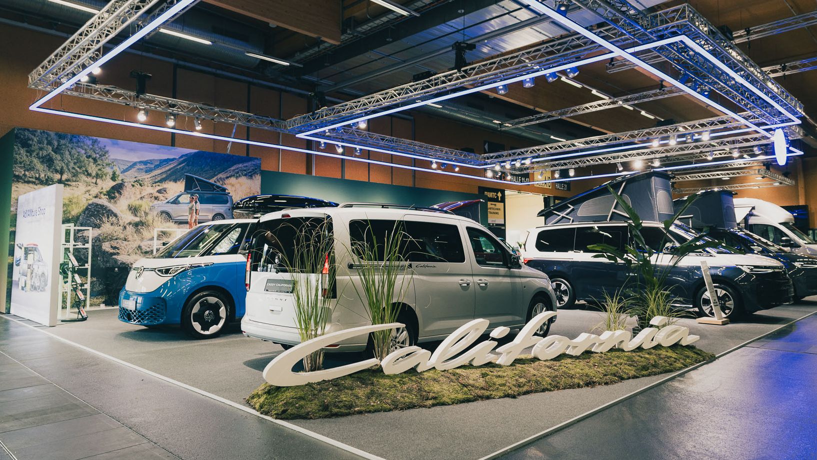 Ein Ausstellungsstand mit verschiedenen Camper-Vans, die unter dem Schriftzug "California" präsentiert werden. Der Bereich ist hell beleuchtet und hat mehrere Pflanzen als Dekoration. Ein großer beleuchteter Rahmen hängt über den Fahrzeugen.