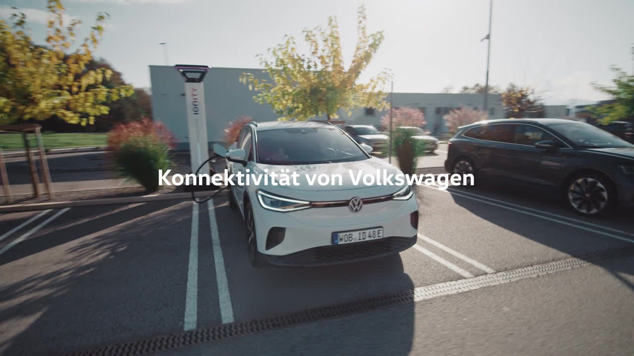 Ein weißer Volkswagen ID.7 steht auf einem Parkplatz neben einer Ladestation für Elektroautos. Im Hintergrund sind weitere Fahrzeuge und moderne Gebäude zu sehen. Text auf dem Bild: "Konnektivität von Volkswagen".