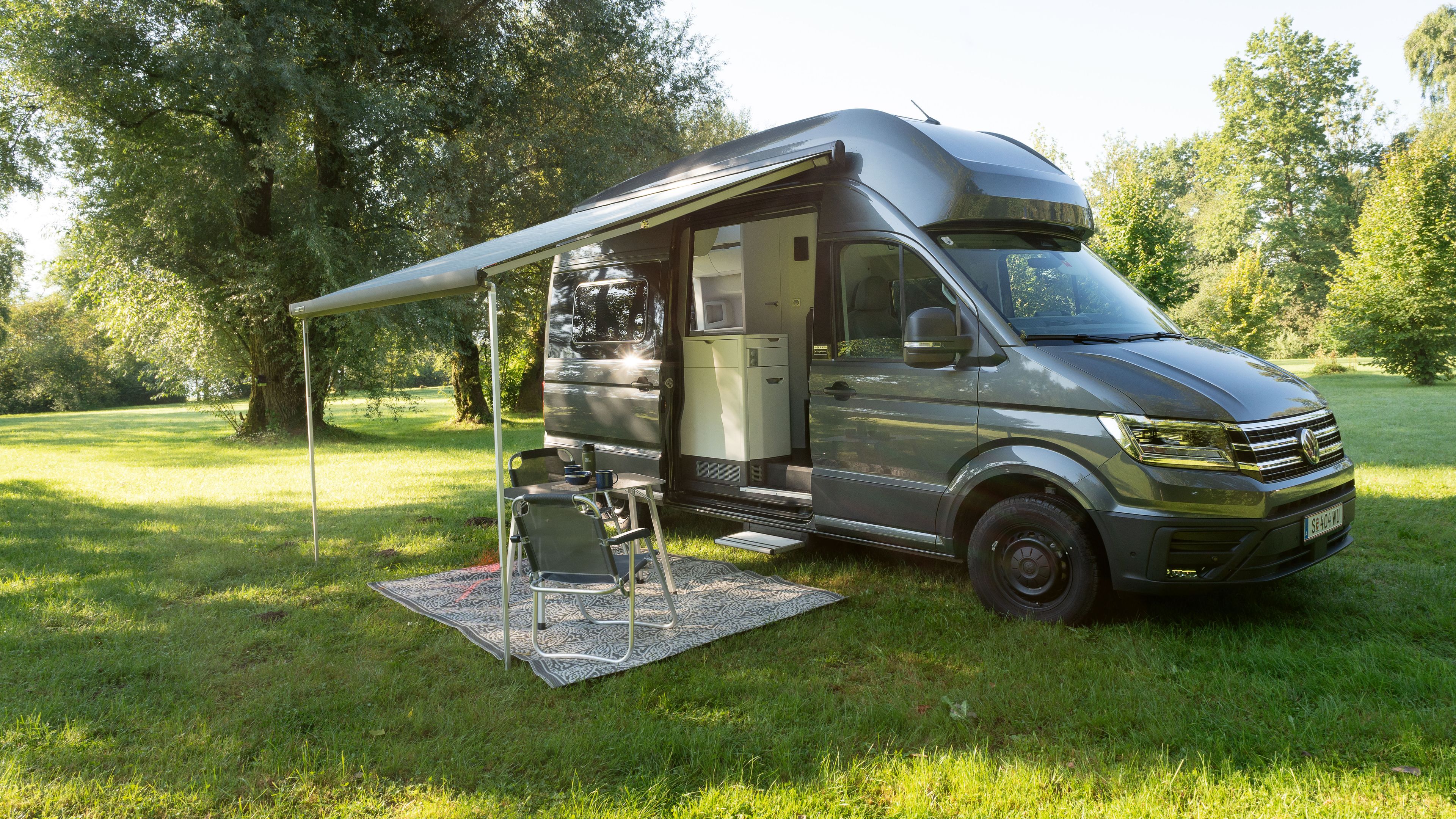 Ein großer grauer Wohnmobil steht auf einer grünen Wiese mit Bäumen im Hintergrund. Das Fahrzeug hat eine ausgefahrene Markise, unter der ein Tisch und zwei Stühle stehen. Im Hintergrund ist eine für Camping geeignete Naturumgebung.