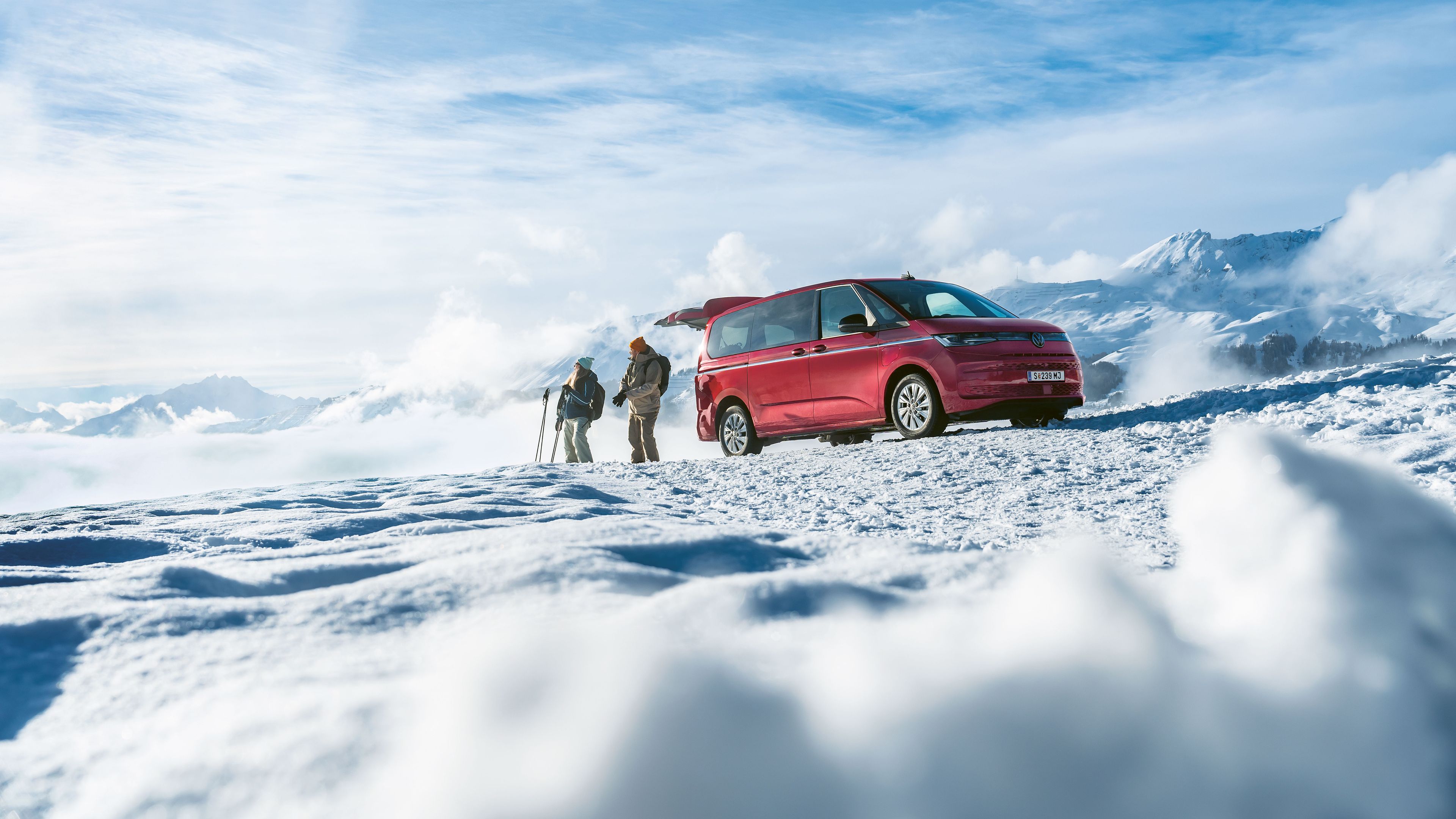 Ein roter Volkswagen Multivan mit geöffneter Heckklappe parkt in den verschneiten Bergen. Ein Mann und eine Frau mit Skiern stehen davor.