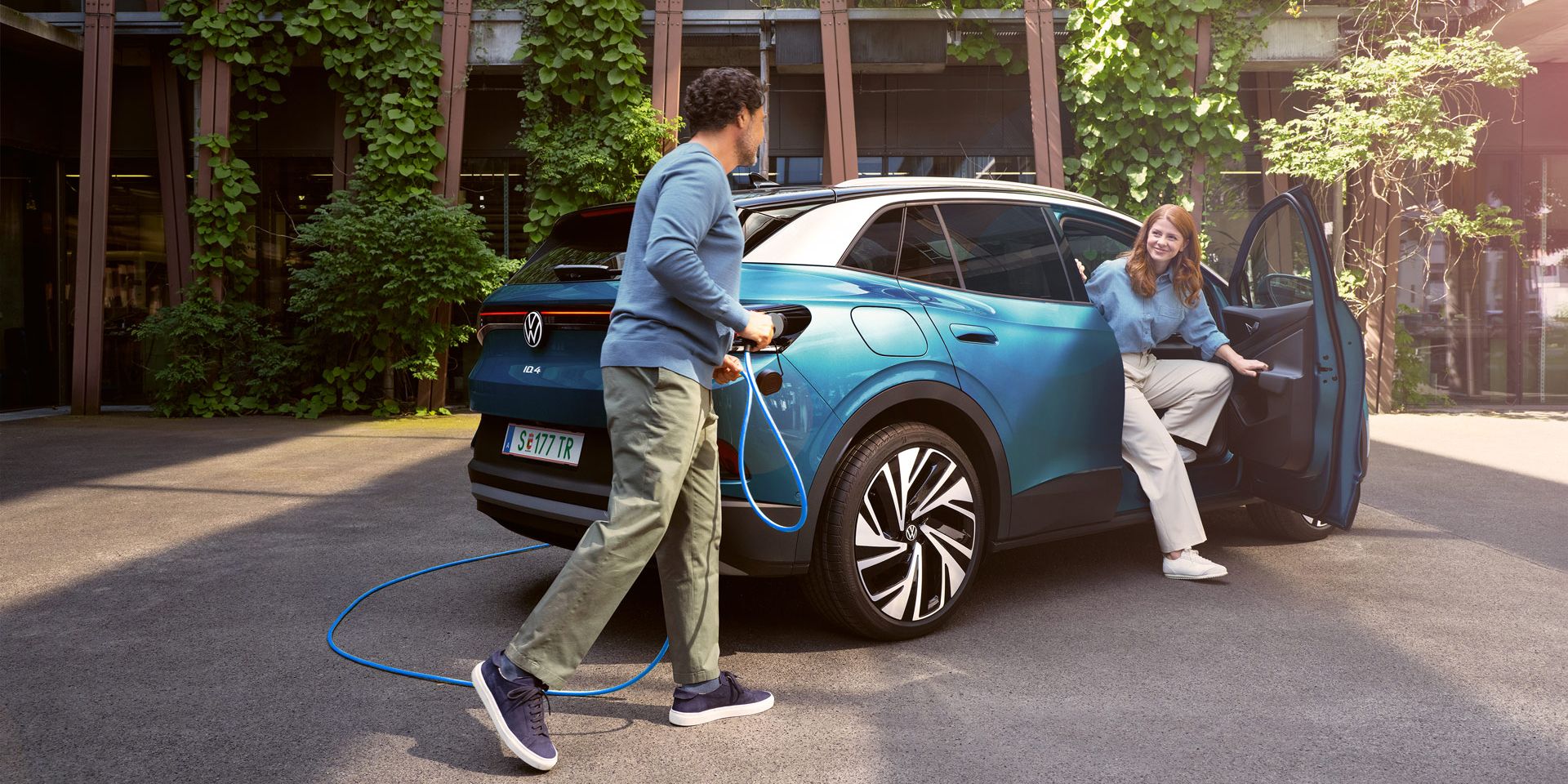 Eine Frau steigt aus einem blauem ID.4 aus, während ein Mann das POWER 2 Go Kabel anschließt. Im Hintergrund sind grüne Pflanzen und moderne Gebäude zu sehen.