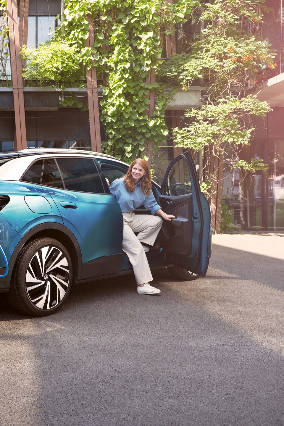 Eine Frau steigt aus einem blauem ID.4 aus, während ein Mann das POWER 2 Go Kabel anschließt. Im Hintergrund sind grüne Pflanzen und moderne Gebäude zu sehen.