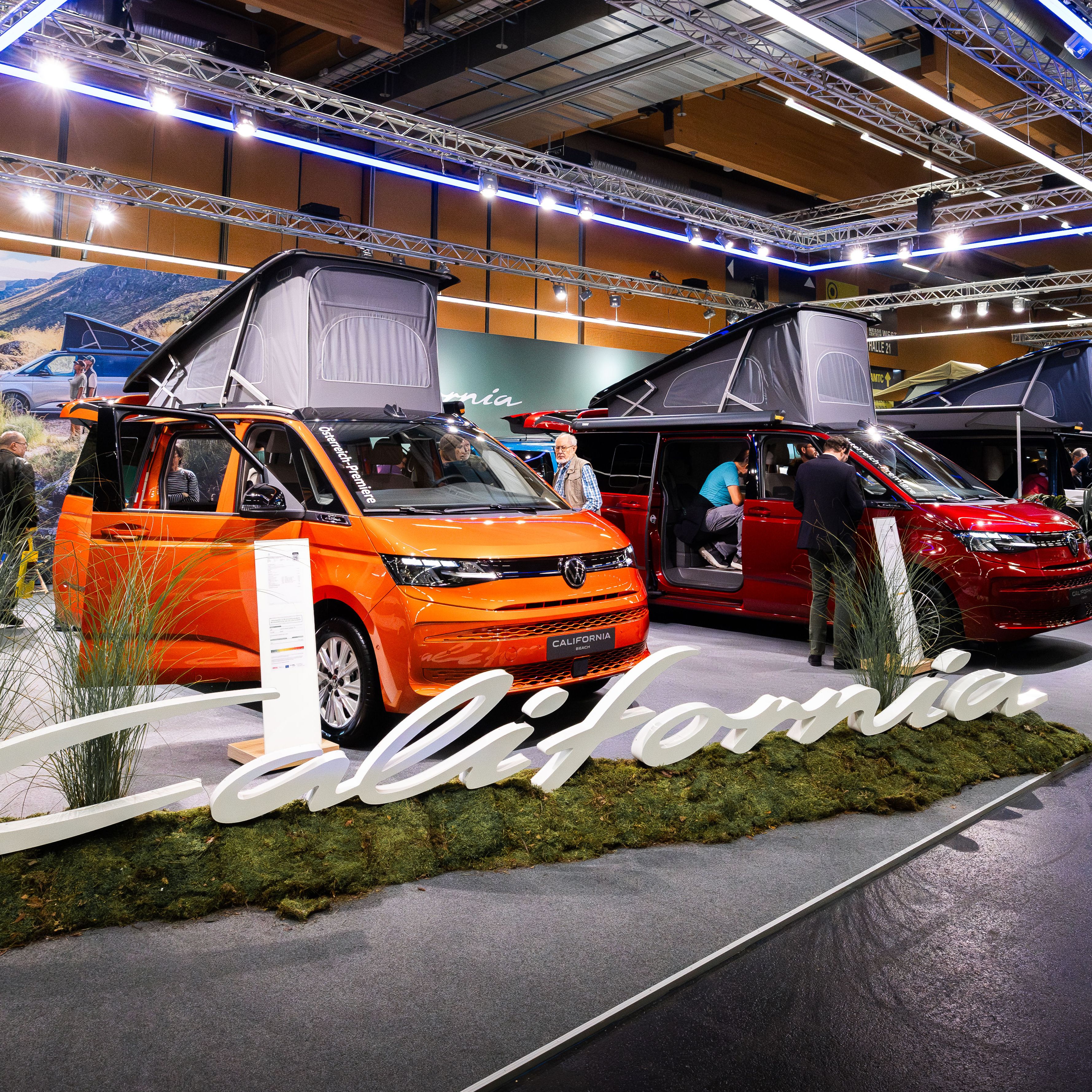 Ein Ausstellungsbereich auf einer Messe zeigt mehrere VW California Modelle. Ein Modell in leuchtendem Orange und ein Modell in Dunkelrot stehen nebeneinander, beide mit ausgefahrenem Dachzelt. Der Bereich ist mit Pflanzen und einem großen "California"-Schriftzug dekoriert.