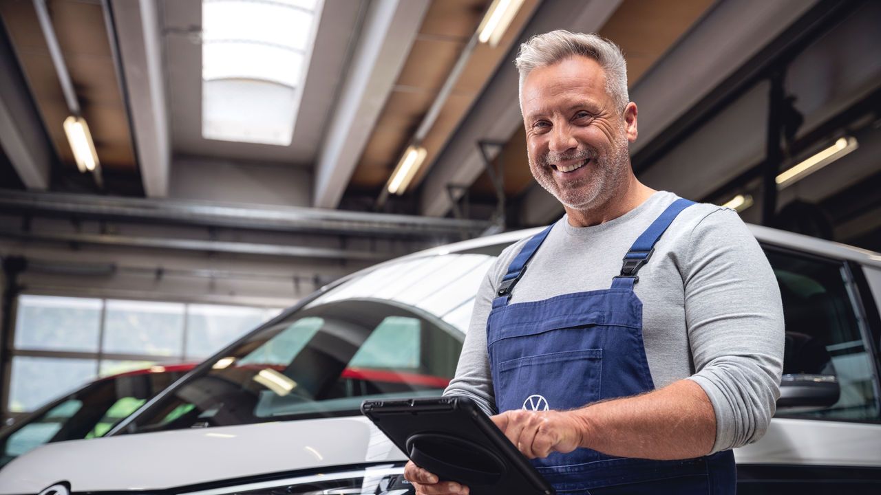 Ein Volkswagen Mechaniker in einer Werkstatt trägt blaue Arbeitskleidung und benutzt ein Tablet. Im Hintergrund steht ein Volkswagen ID. Buzz.