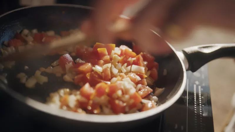 Eine Person kocht gehacktes Gemüse wie Paprika und Zwiebeln in einer Pfanne. Die Zutaten werden bei mittlerer Hitze angebraten. Die Hand der Person rührt das Gemüse mit einem Kochlöffel um.