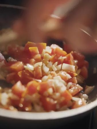 Eine Person kocht gehacktes Gemüse wie Paprika und Zwiebeln in einer Pfanne. Die Zutaten werden bei mittlerer Hitze angebraten. Die Hand der Person rührt das Gemüse mit einem Kochlöffel um.