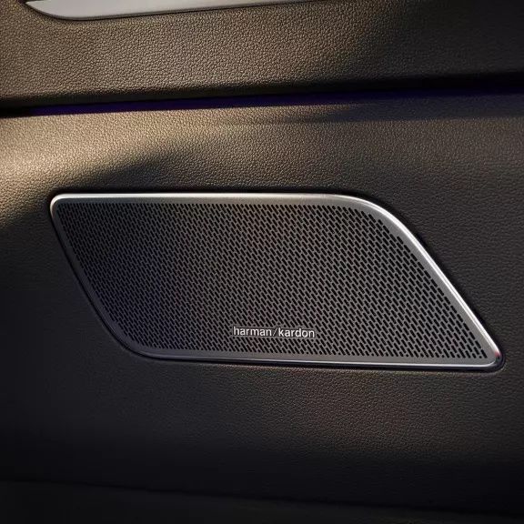 Detailaufnahme einer Autotürverkleidung mit einem integrierten Lautsprecher. Der Lautsprecher ist rechteckig mit abgerundeten Kanten, von einem silbernen Rahmen umgeben und trägt das Logo „Harman Kardon“. Die Türverkleidung ist dunkel und wirkt hochwertig verarbeitet.