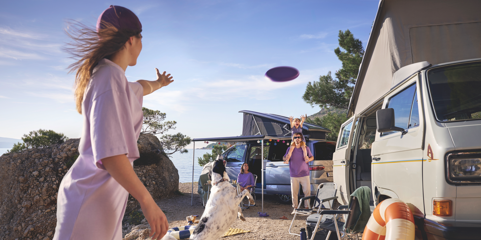 Eine Familie spielt auf einem Campingplatz, im Hintergrund ist ein Volkswagen California zu sehen.
